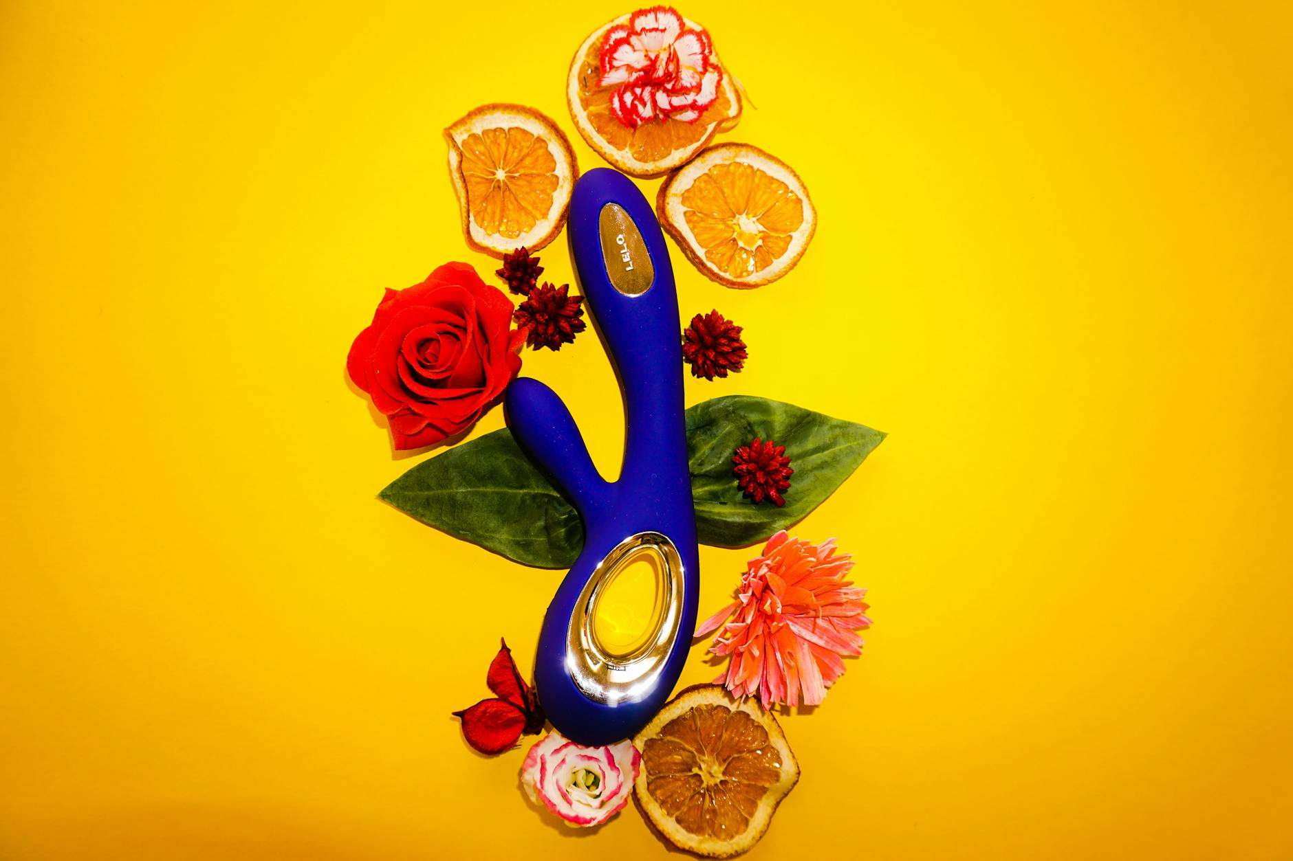 Vibrant blue vibrator surrounded by flowers and citrus on yellow background